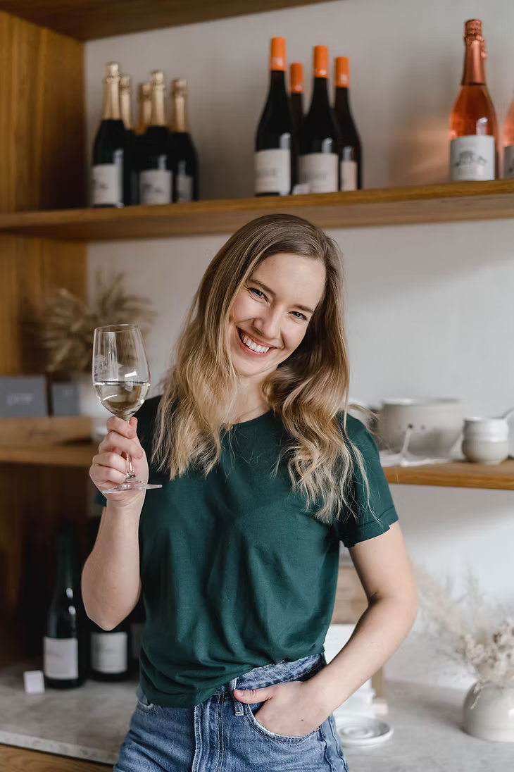 Frau mit einem Glas Wein in der rechten Hand mit verschiedenen Weinen und Keramikstücken im Hintergrund. 