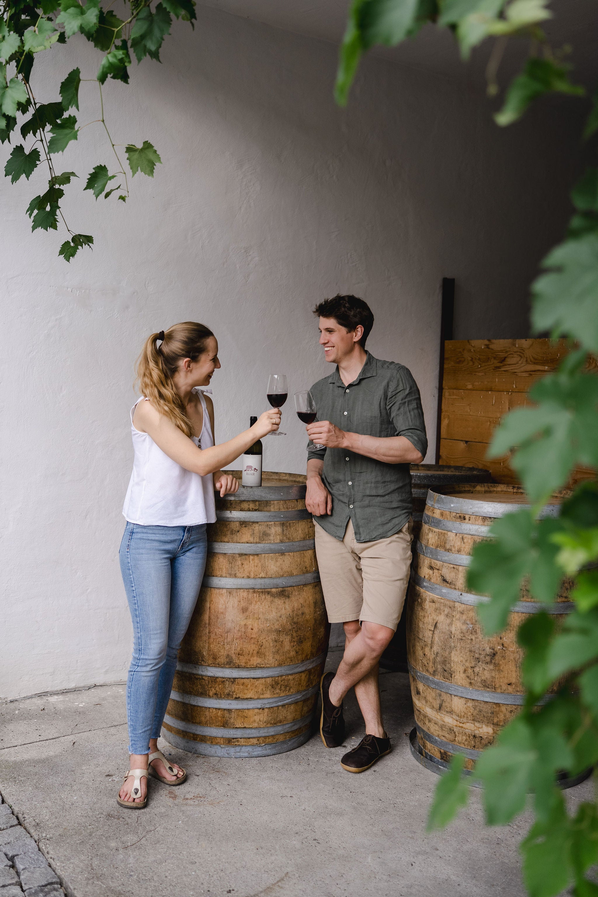 Frau und Mann lehnen an Weinfass und stoßen mit einem Glas Rotwein an. 
