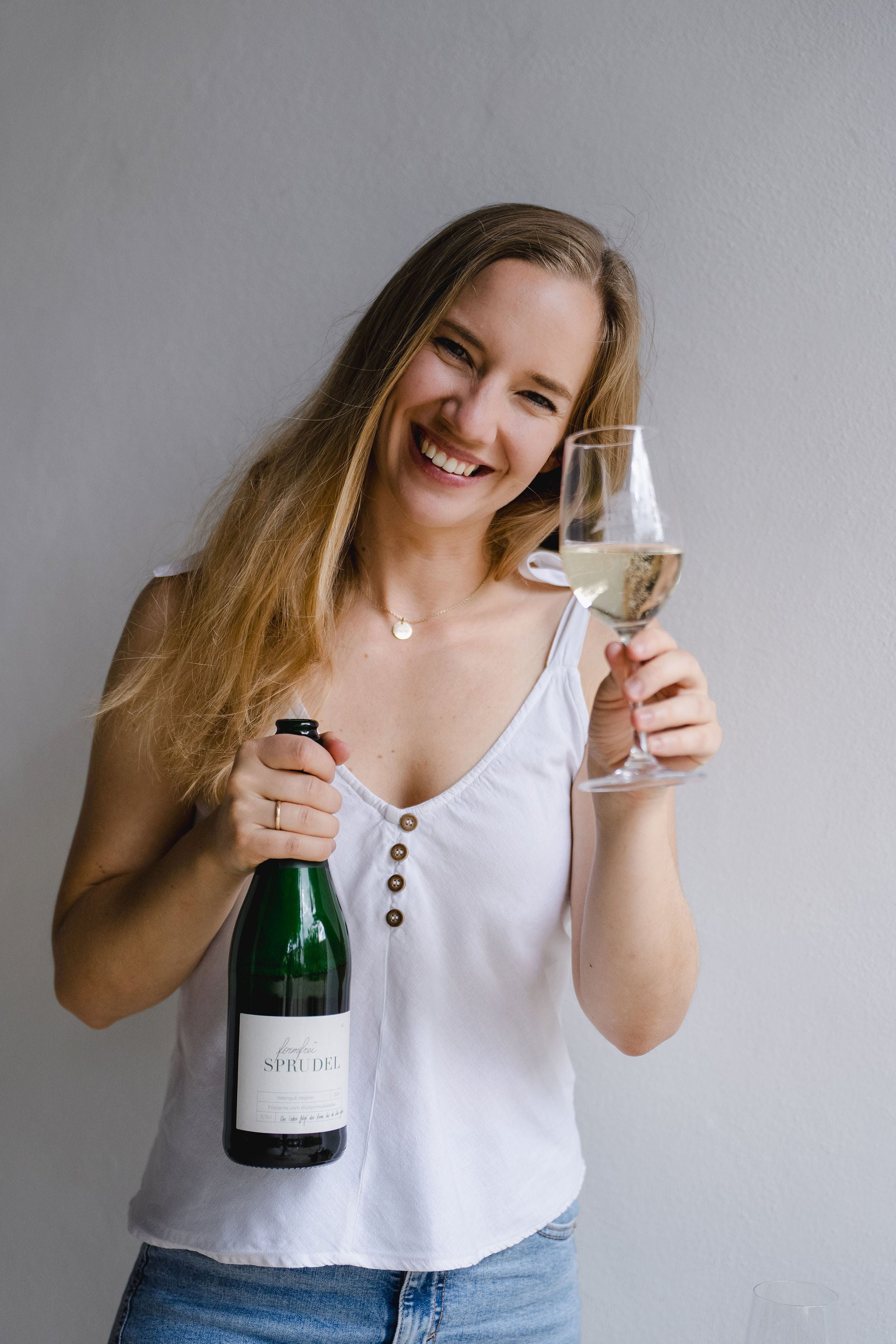 Frau hält formfrei Sprudel Flasche in der rechten und ein gefülltes Weinglas in der linken hand und lacht in die Kamera 